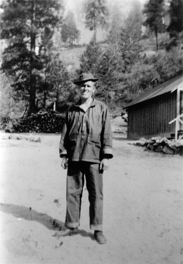 A CCC man stands in the CCC Camp, a firewood pile is in the background. Writing beneath the photo reads: '1850.0016. Modeling CCC fatigue work wear at Camp S-223, McCall'