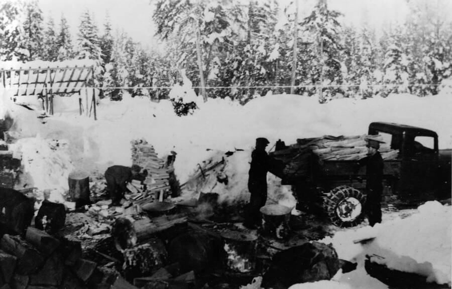 Three CCC men cutting firewood in the snow and loading firewood into a truck. Writing beneath the photo reads: '1850.0018. Firewood cutters, S-223, McCall, 1935'.