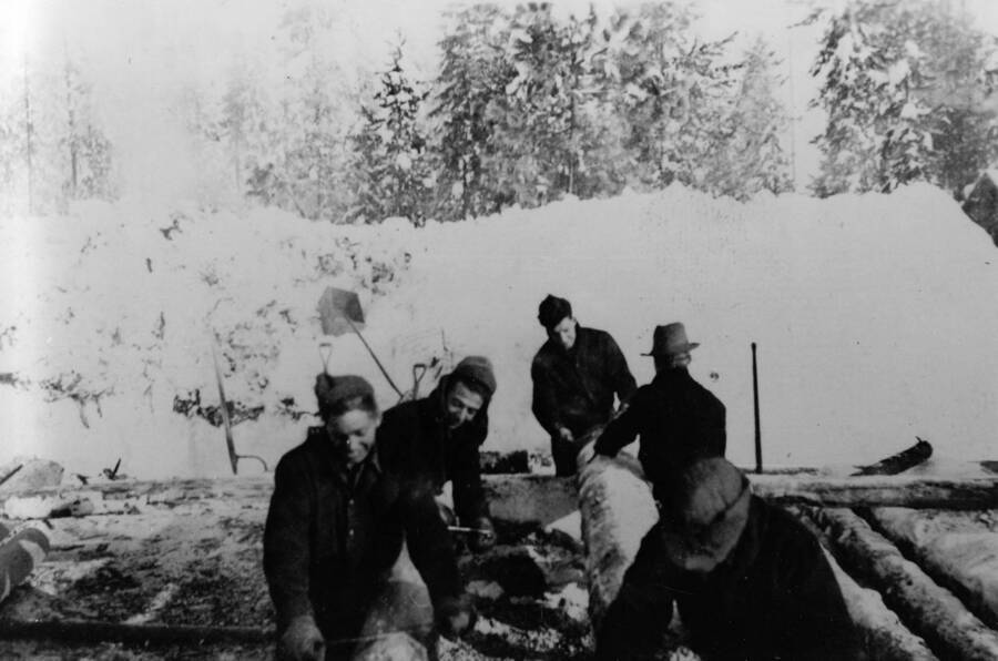 A crew of CCC men working in the snow, carrying logs. Writing beneath the photo reads: '1850.0019. Wood crew, S223, McCall,1935'.