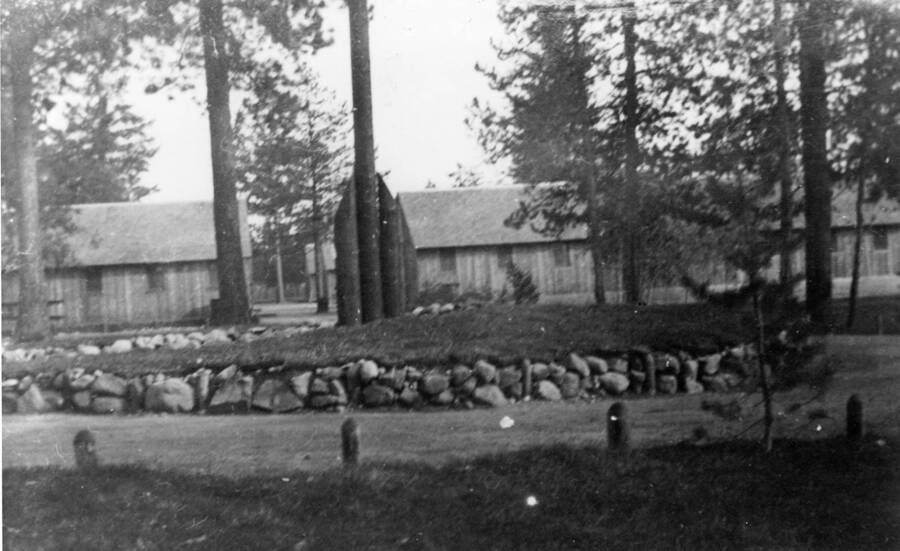 A circle of stone surrounds a small hill with a flagpole in the center at CCC Camp S-223. Writing on the photo reads: '1850.0026. Camp Circle with flag pole and Barracks in the background, S-223, McCall, 1935'.