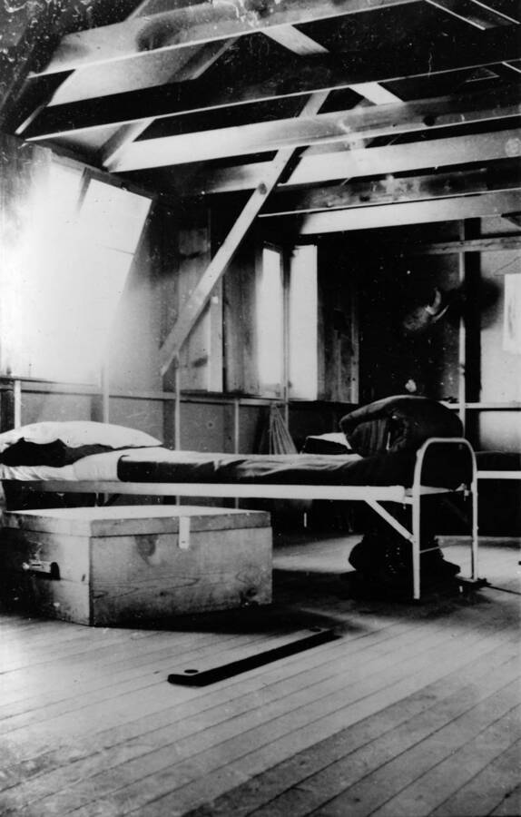 Interior of barrack #5 showing a cot and wooden trunk beneath a window. Writing beneath the photo reads: '1850.0028. Interior of Barracks #5, S-223, McCall, 1935'.