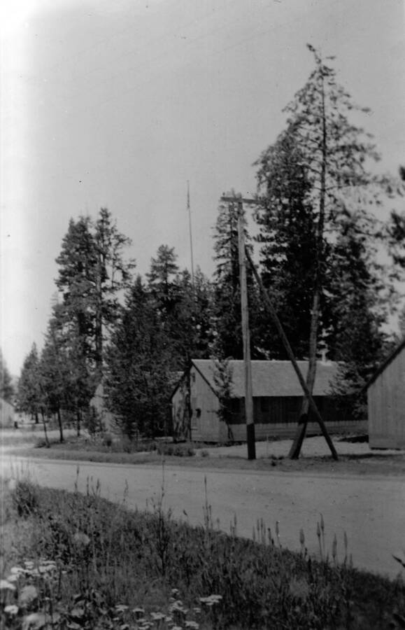 Two barracks can be seen across the road, a utility pole and line of trees in front of them. Wildflowers can be seen in the foreground. Writing beneath the photo reads: 'Barracks 2, 4, and 6 from Front Street, S-223, McCall, 1935.'