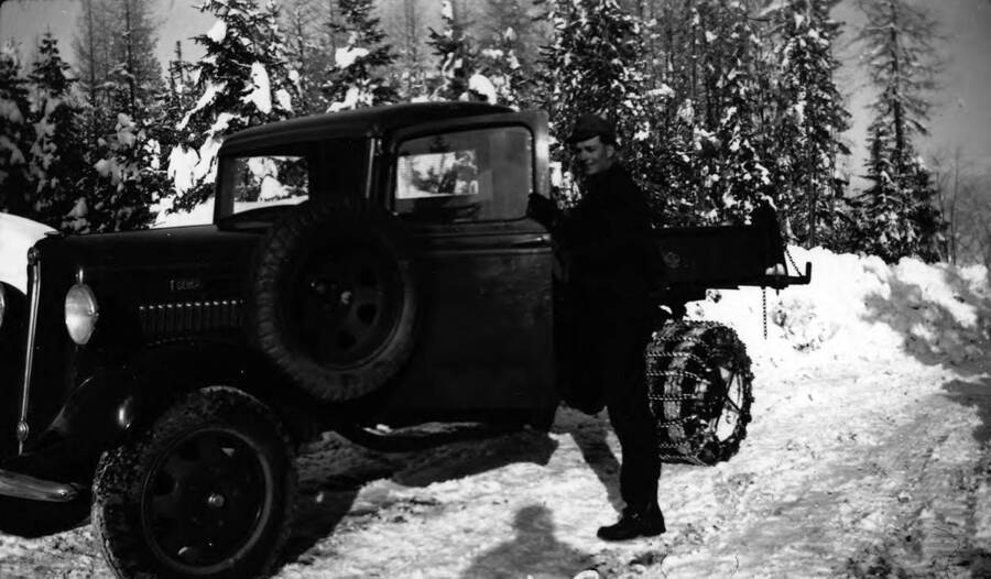 A CCC man poses before entering a truck parked on a snowy road with trees in the background. Writing beneath the photo reads: '1850.0039. CCC member and assigned truck, S-223, McCall, 1935-36'.