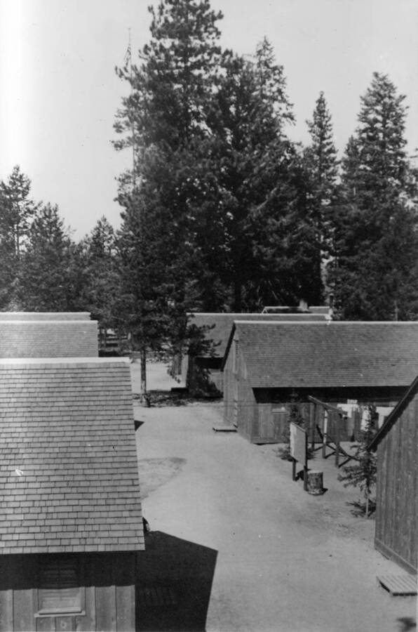 Two rows of barracks viewed from above. Trees are scattered between the buildings and there is a bulletin board in the center of the right row of barracks. Writing beneath the photo reads: '1850.0047. Company street, S-223, McCall, 1935'