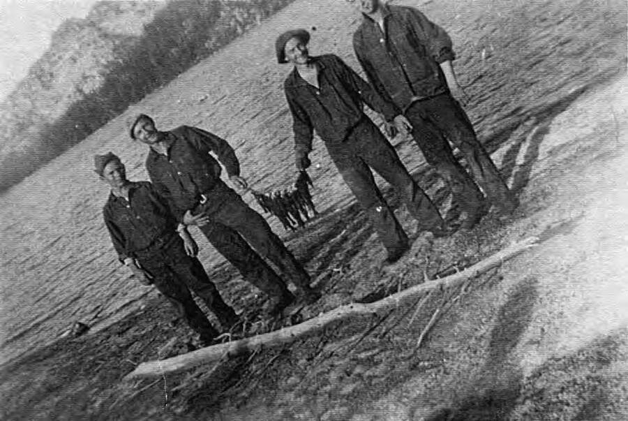 Four men stand holding a line of fish caught from Lake Cascade at Camp French Creek. Writing beneath the photo reads 'Photo No. 1850.0051. Fish on line hooks, Camp French Creek, 1939.'