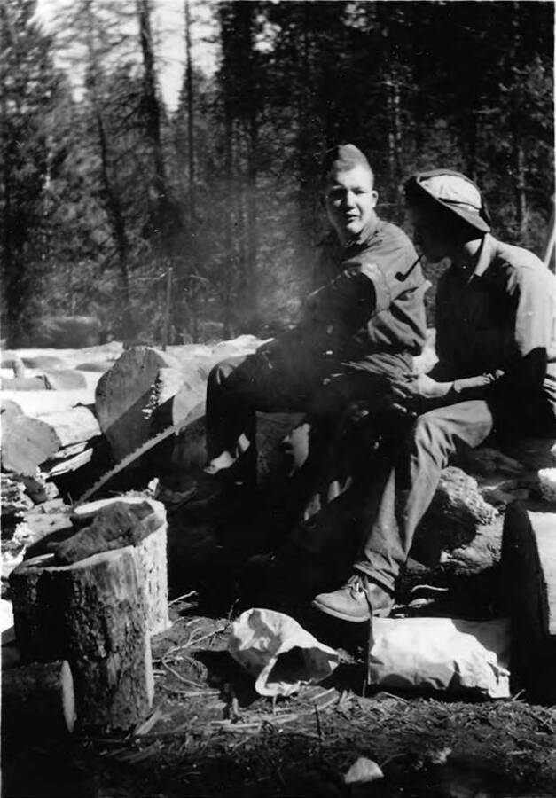 Two men sit and eat their lunches. Empty paper bags can be seen on the ground. They are sitting on cut logs. Writing beneath the photo reads 'Photo No. 1850.0057. 'Tarzan 'cat-skinner' of the 'destruction gang,' C-1997 (?)'