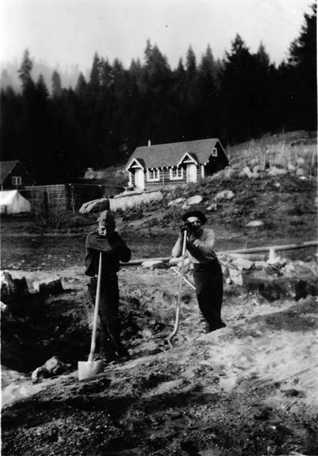 Two men lean on shovels outside of the Company's house. Writing beneath the photo reads: 'Photo No. 1850.0077. Men leaning on shovels, 'Hard at work, Co's house (C-1997 ?)'