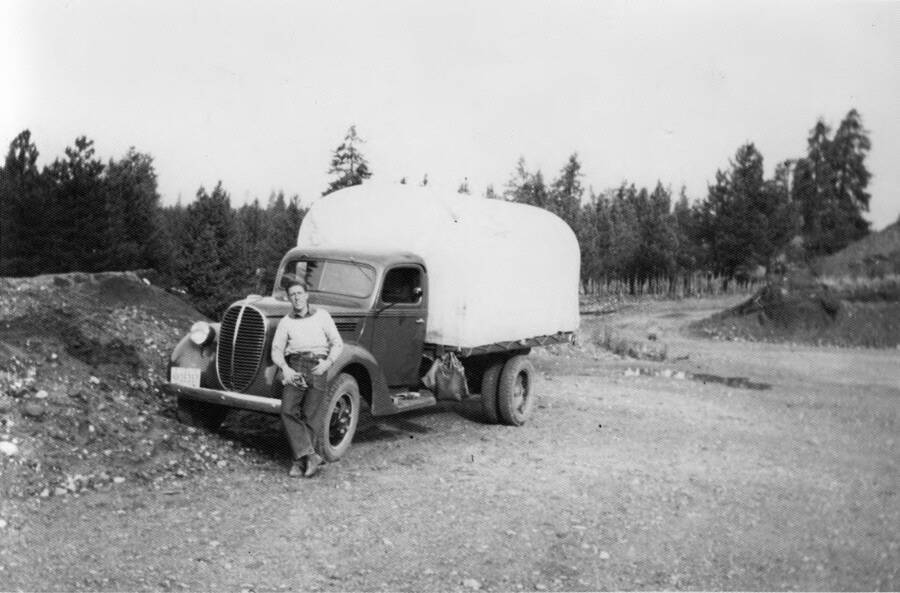 A man stands in front of a 1937 Ford Truck that serves as one of the camp's trasnport vehicles. The truck has a covered load tied to the flatbed of the truck. Writing beneath the photo reads: 'Photo No. 1850.0078. Transport and driver, Smith's Ferry, C-1997 (?)'