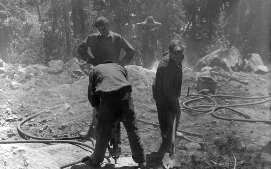 Five men work at a project site. One man is digging with a shovel and another spots while one man operates a pneumatic jack hammer. Writing beneath the photo reads: 'Photo No. 1850.0088. View of CCC work, South Fork Road, 1939.'