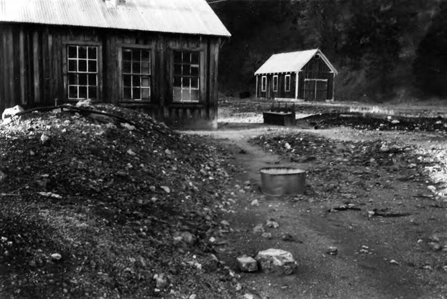 Two buildings at the Smiths Ferry camp. A fire pit can be seen in the foreground near the back of the first building. Writing beneath the photo reads: 'Photo No. 1850.0091. Smiths Ferry, buildings, C-1997'