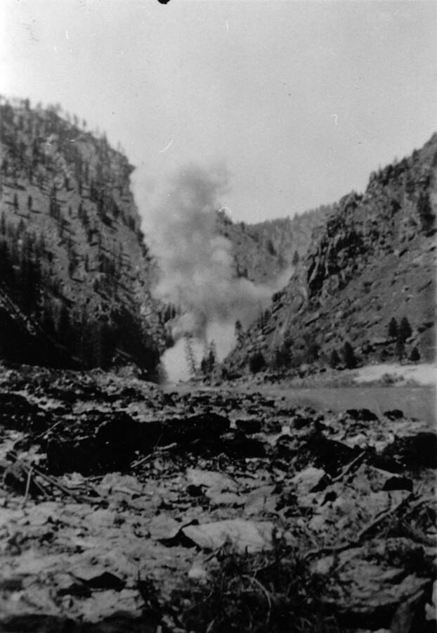 Road blasting and bridge foundation construction can be seen in the distance during the winter in the Idaho National Forest. Writing beneath the photo reads: 'Photo No. 1850.0097. Blasting road and bridge foundation, Idaho National Forest, F-108, 1935-36.'