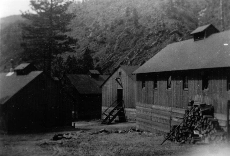 A photograph of four buildings at Camp French Creek, with wood stacked behind the buildings. Photo No. 1850.0098. Camp French Creek, F-108, Idaho National Forest, 1935-36