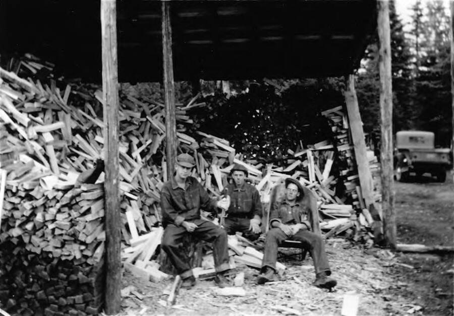 Three men rest near a wood pile at one of the camp project sites. One man appears to be asleep in a chair, while the other two sit on wood stacked in piles. Writing beneath the photo reads: 'Photo No. 1850.0087. Men resting near wood pile, captioned 'Industry,' F-414, C-1997 (?).'
