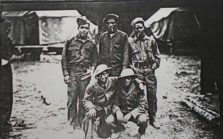 Group photo of a few African American enrollees at a CCC Camp in Northern Idaho. The men can be seen wearing hats and standing and squatting with their arms wrapped around each other.