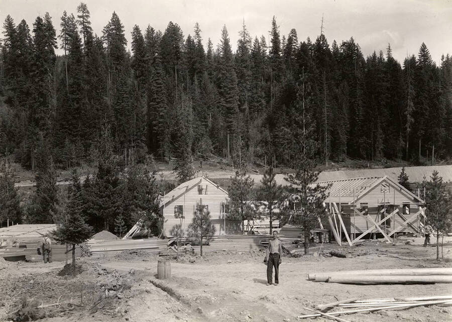 View of three buildings under construction at Hayden Creek CCC Camp. One construction worker stands in the foreground.