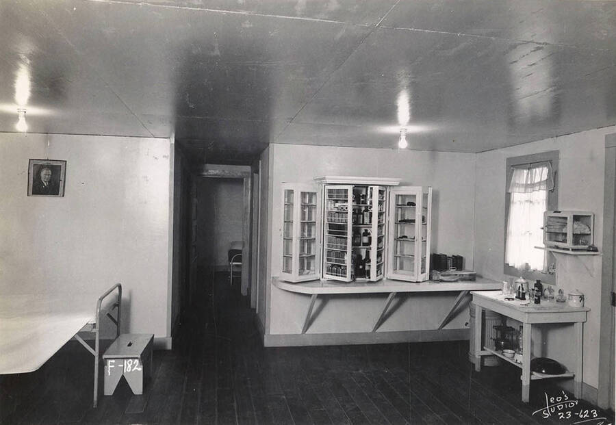 Interior view of an infirmary at Wolf Lodge Bay CCC Camp, F-182. Note the cabinet filled with medicine bottles.  Writing on the bench reads: 'F-182'. Back of photo reads: 'Wolf Lodge Beauty Bay'.
