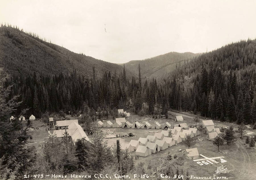 A view of Horse Heaven CCC Camp. On the right side of the photo is a geoglyph or 'hillside letters' displaying F-156. Note a half-built tent frame in the center of the clearing. Writing on the photo reads: '21-473 Horse Heaven CCC Camp F-156 Company 569 Enaville, Idaho. Leo's Studio Spokane'.