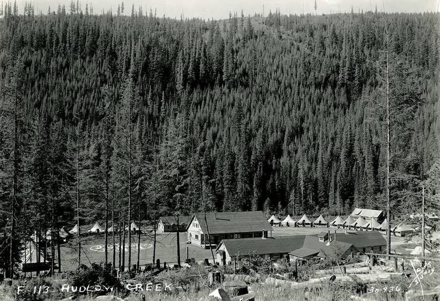 A view of Hudlow Creek CCC Camp. In the center of the clearing, through the trees is an upside-down geoglyph that reads: 'F-113 [546] CCC'. Writing on the photo reads: 'F-113 Hudlow Creek Leo's Studio.'