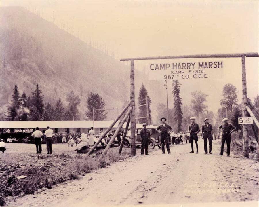 Prichard Camp entrance with sign and personnel posing on the road. Camp F-30, company 967, was renamed Harry Marsh Camp. Writing on the sign reads: 'Camp Harry Marsh (Camp F-30) 967th Company CCC'. The small sing on the right of the photo reads: '5 miles per hour'. There also appears to be a baseball game occurring in the background.