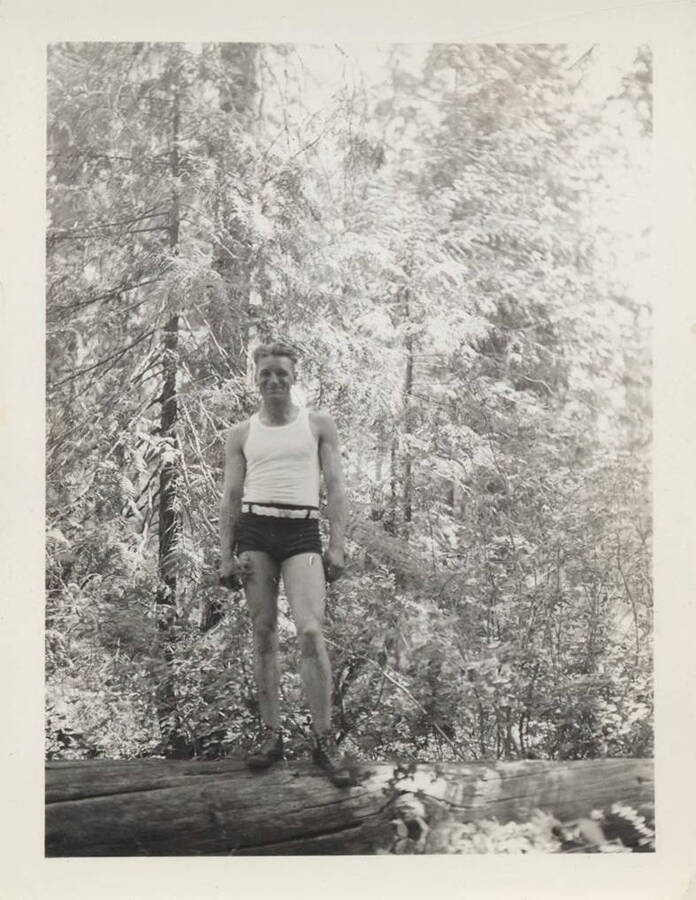 Camp Superintendent H. E. Matthews of Camp F-139 stands on a downed log wearing a tank top and shorts and hiking boots.