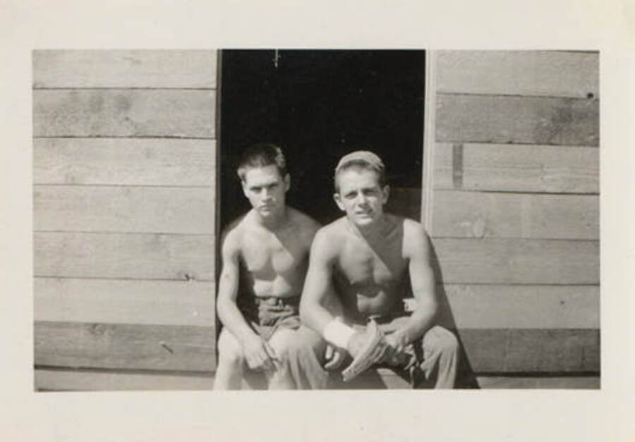 Two shirtless enrollees sit in the doorway of a building. The back of the photograph reads: 'just two mugs, '