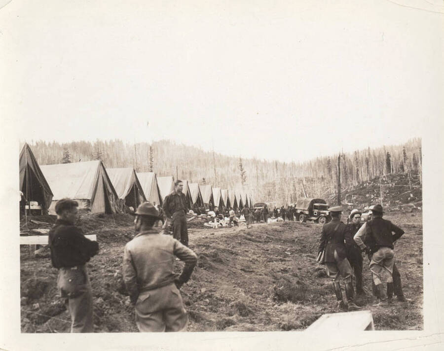 Two enrollees talk in the foreground while another group of enrollees stands to the side upon arrival at Camp Prichard. The back of the photograph reads: 'The camp as it was the day we arrived.'