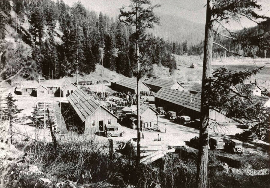 Overview of Cataldo CCC Camp. CCC men are gathered around the flagpole. Back of photo reads: 'Gene Fuson 773-5420 1933 (or 1937) Cataldo, Idaho'.
