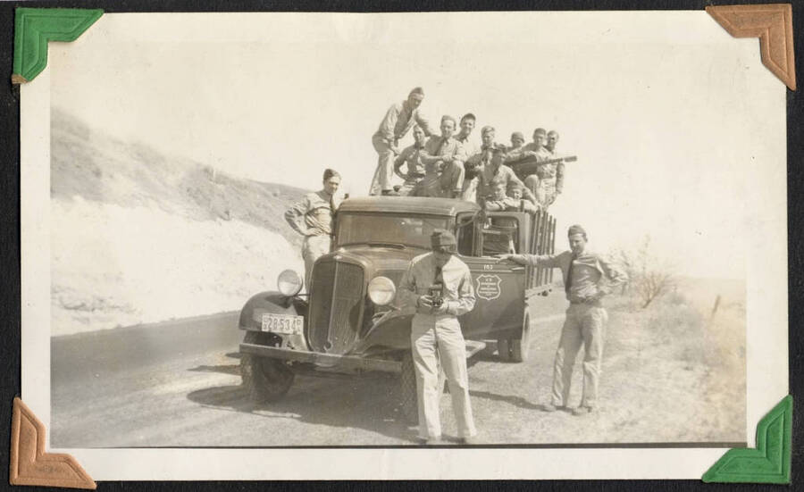 Men in uniform loaded onto a truck, one holding a guitar and one in front with a handheld camera. SCS-1, C-1503. From the Paul Saft photographic album, SCS-1, C-1503, 1938-39, depicting camp life, taken mostly in the Moscow, Lewiston, Robinson Lake areas.