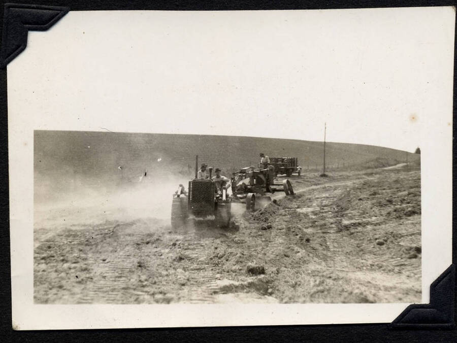 Work crew with tractor pulling a grader leveling a road surrounded by fields. SCS-01, C-1503. From the Paul Saft photographic album, SCS-1, C-1503, 1938-39, depicting camp life, taken mostly in the Moscow, Lewiston, Robinson Lake areas.
