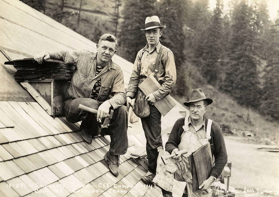 Three CCC men posing for a picture on a roof with shingle hammers and other roofing materials at Cataldo CCC Camp. Writing on the photo reads: 'Shinglers Three CCC Camp F-114 Company 967 Cataldo, Idaho Photo by Leo's Studio Spokane'.