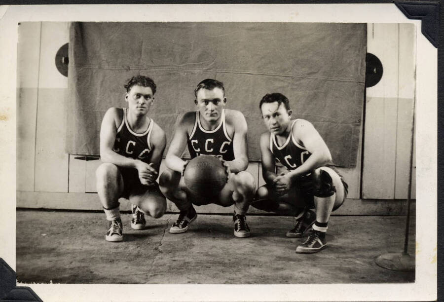 Three kneeling basketball players in CCC jerseys. SCS-1, C-1503. From the Paul Saft photographic album, SCS-1, C-1503, 1938-39, depicting camp life, taken mostly in the Moscow, Lewiston, Robinson Lake areas.