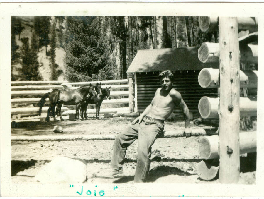 Picture from album of enrollee sitting on poll of corral with two horses in background. Krassel. Handwritten caption ''Joie''.  This is likely to be located at Camp Creek, South Fork of the Salmon River, which built Krassel Ranger Station.
