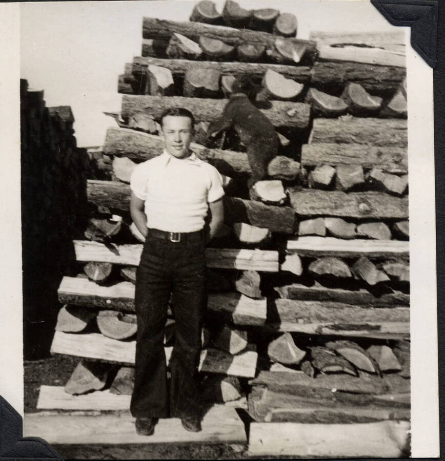 Man posed in front of large wood pile, with bear cub on chain in the background. SCS-1, C-1503. From the Paul Saft photographic album, SCS-1, C-1503, 1938-39, depicting camp life, taken mostly in the Moscow, Lewiston, Robinson Lake areas.