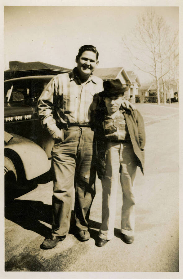 Two men stand in the road and in front of a street filled with houses and a truck. Back of photograph reads, "John + Shine."