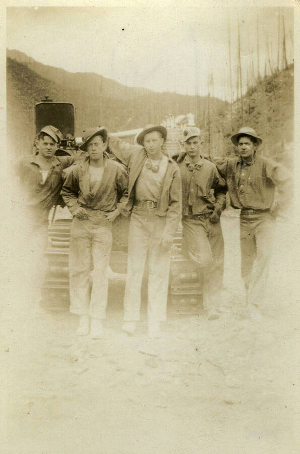 Five men wearing work wear and hats pose in front of a car with a landscape of barren trees and mountains in the background. Back of photograph reads, "Jack hammer crew North Fork."