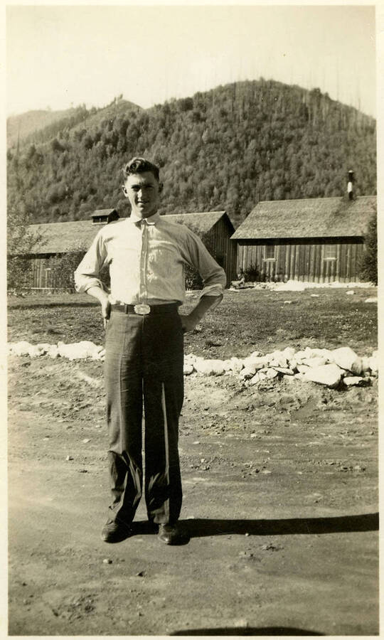 A man standing in front of two buildings and a mountainous landscape in the background. Back of photograph reads, "Madkins CCC camp."