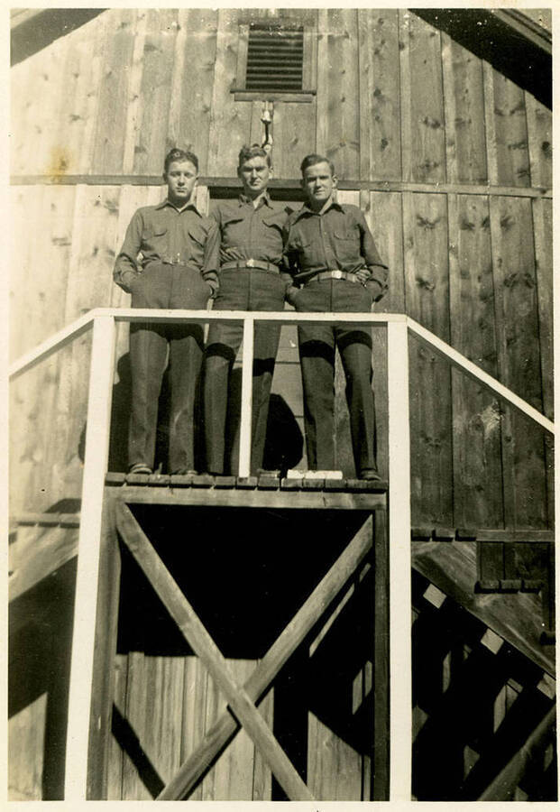 Three men dressed in uniform pose for a photograph on the top of a flight of stairs and in front of a wooden building. Back of photograph reads, "Arnold was in CCC 1936-1937 3 friends" and "Three Rookies at North Fork."