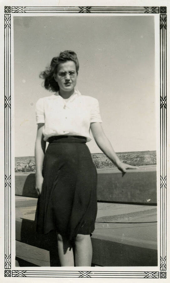 A woman wearing glasses poses on what looks like a bridge over a body of water. Back of photograph reads, "Friend."