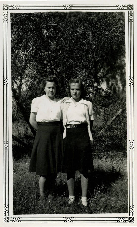 Two women wearing skirts pose in front a tree and grassy landscape.