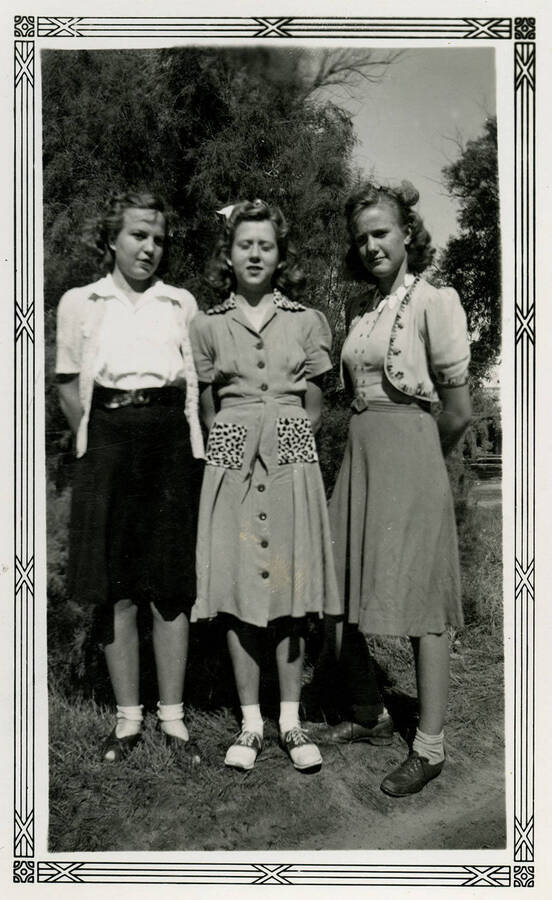 Three women wearing dresses and skirts stand in front of a tree. The woman in the middle closes her eyes.