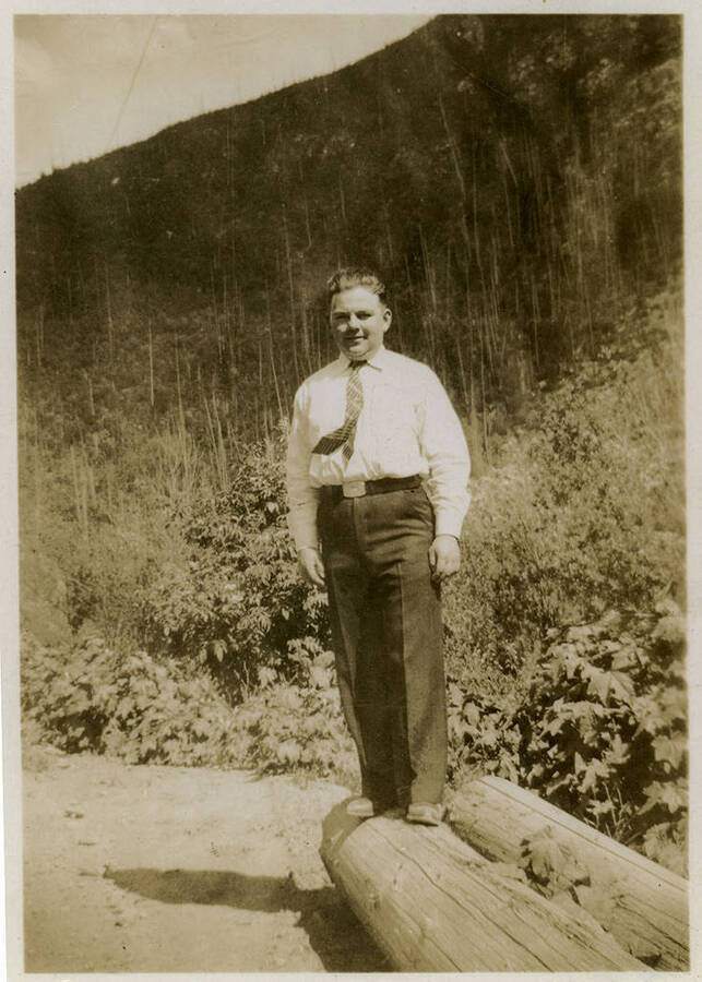 A man wearing a tie stands on a log in front of a mountainous landscape. Back of photograph reads, "Sims. CCC Co. 5702."