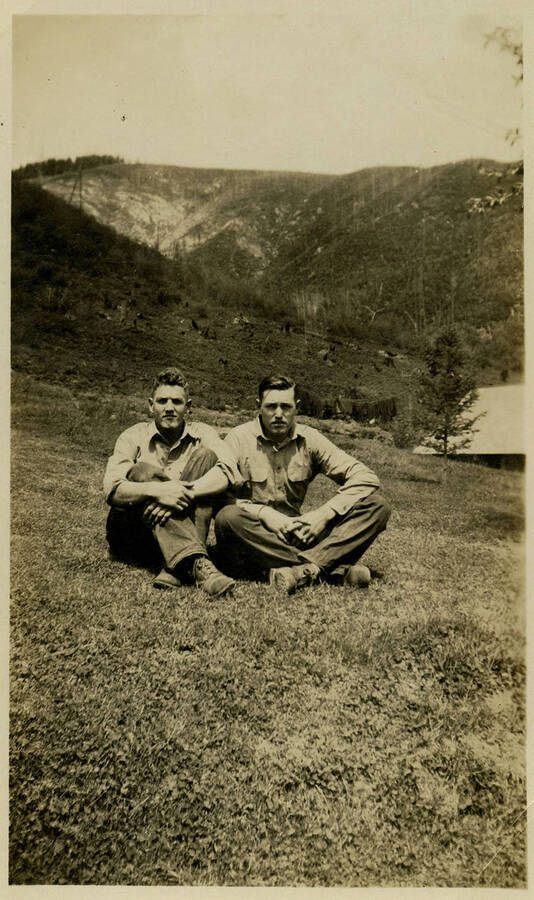 Two men wearing similar clothes sit on a grassy field in front of a mountainous landscape. Back of photograph reads, "Wealy + Brown North Fork Idaho."