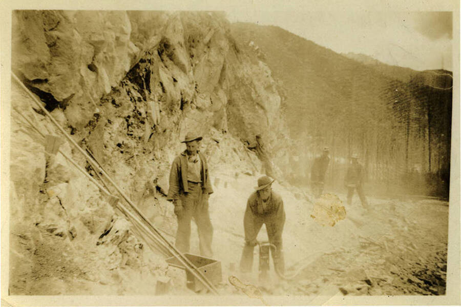 Two men in the foreground stand near a rocky formation, the man on the right is bent and holding a jack hammer. Three other figures can be seen in the background. Back of photograph reads, "Rookies at North Fork."