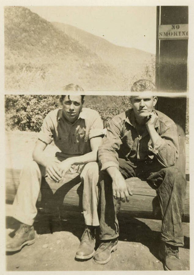 Two men sitting on a log. A sign reads, "No smoking" on a pillar in the background. Mountains can also be seen in the background. Back of photograph reads, "Truck driver and Truck master North Fork."
