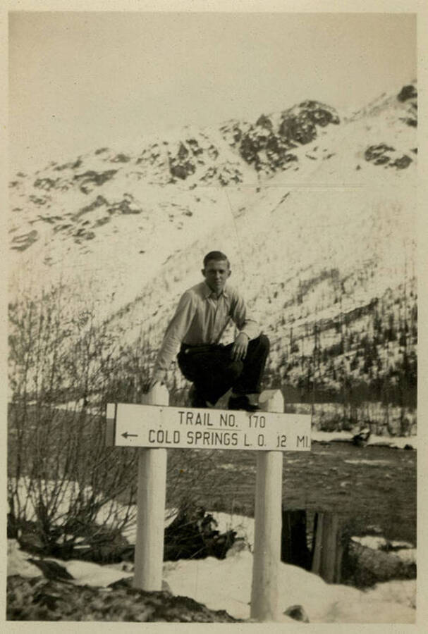 A man, possibly Norris, sitting on a sign that reads, "Trail No. 170 Cold Springs L.O. 12 Mi." Snowy mountains can be seen in the background. Back of photograph reads, "Norris CCC. Co. 5702."