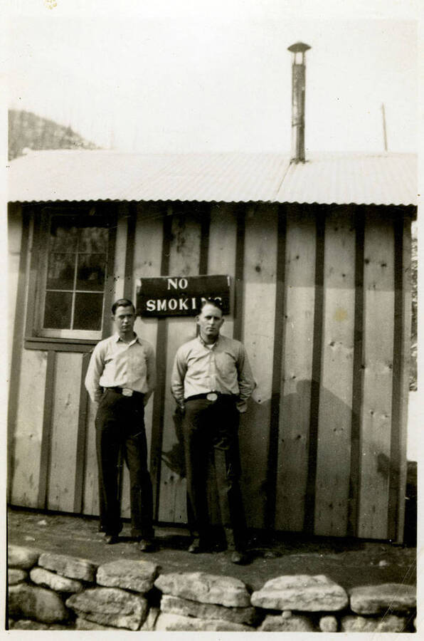Two men wearing similar clothes stand in front of a building and a sign that reads, "No Smoking." Back of photograph reads, "Herric + Norris North Fork."