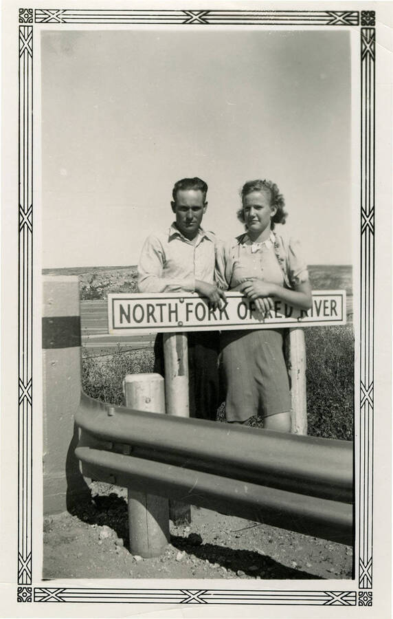A man and woman stand behind a sign that reads, "North Fork of Red River." Back of photograph reads, "Friends."