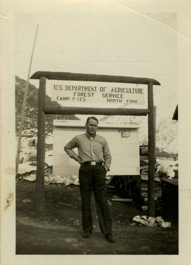 A man stands under an outdoor sign that reads "U.S. Department of Agriculture Forest Service Camp F-123, North Fork." Back of photograph reads, "Herric Camp North Fork."
