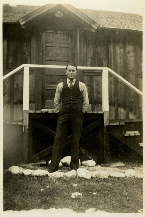A man, possibly Herric, stands in front of a wooden building. He wears a waistcoat and tie. Back of photograph reads, "Herric North Fork."