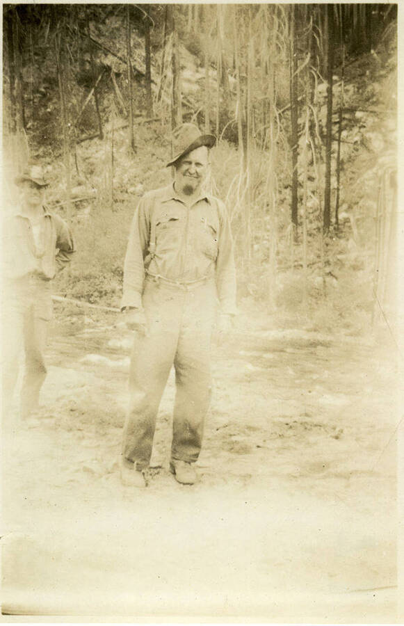 The foreman of jack hammer crew, (possibly Mr. Ryan) stands in front of a landscape of bare trees. Another man can be seen in the left background. Back of photograph reads, "Foreman of Jack hammer crew North Fork, Mr. Ryan."